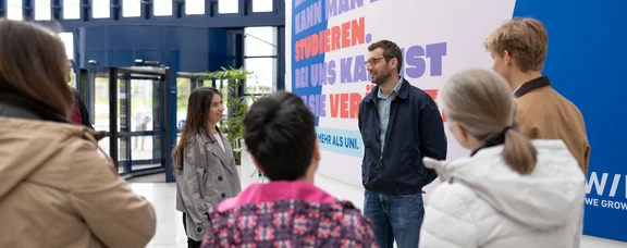Mach dir mit unseren Infoangeboten ein Bild von der Uni Witten/Herdecke. Eine Gruppe Personen steht vor einem Plakat.