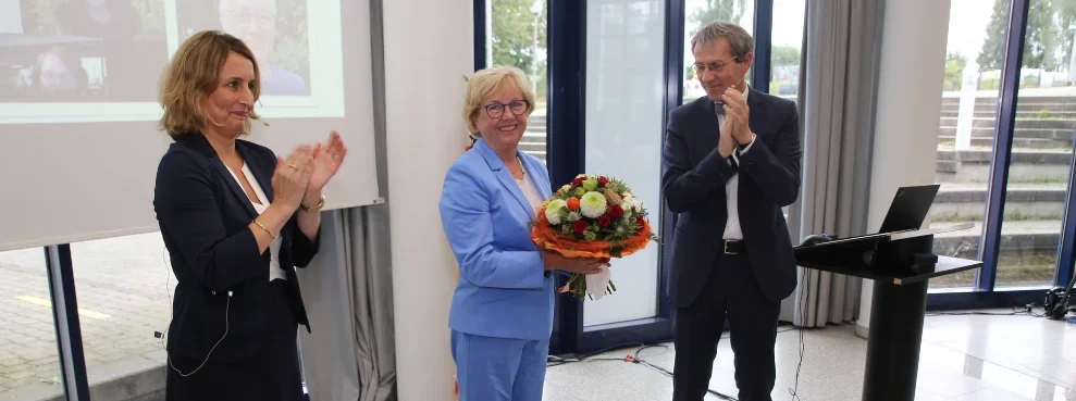 Drei Personen stehen in einer Reihe, die Frau links und der Mann rechts klatschen, die Frau in der Mitte hält einen Blumenstrauß in der Hand.