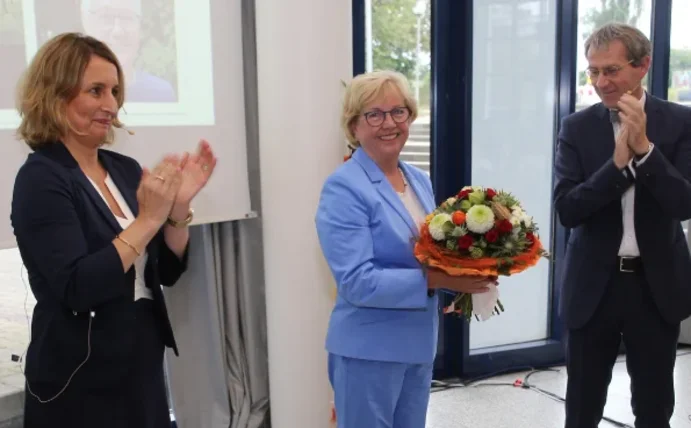 Drei Personen stehen in einer Reihe, die Frau links und der Mann rechts klatschen, die Frau in der Mitte hält einen Blumenstrauß in der Hand.