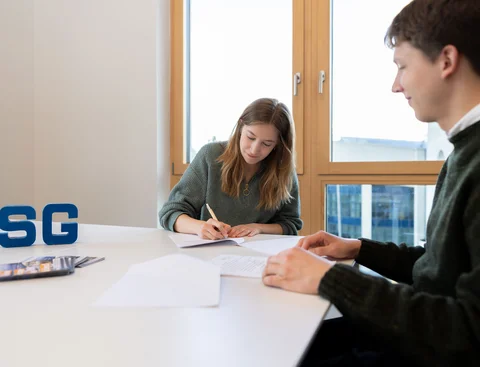 Beratung durch die StudierendenGesellschaft (Foto: UW/H | Michael Schwettmann) Beratungsgespräch zwischen einen Mann und einer Frau. Die Frau unterschreibt einen Vertrag. Auf dem Tisch zwischen ihnen stehen die Logo-Buchstaben der StudierendenGesellschaft (SG).