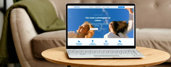 Ein Laptop steht auf einem Couchtisch. Im Hintergrund sind ein Sofa und eine Wolldecke.