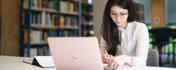 Arbeitsplatz in der Bibliothek (Foto: UW/H | Johannes Buldmann) Eine Studentin sitzt an einem Schreibtisch und lernt. Vor ihr steht ein Laptop, daneben liegt ein aufgeschlagenes Buch. Im Hintergrund stehen Bücherregale.