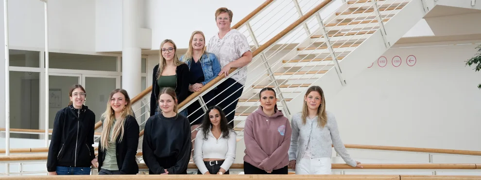 Gruppenfoto von neun Personen, die teils in einer Reihe, teils auf einer Treppe im Hintergrund stehen.