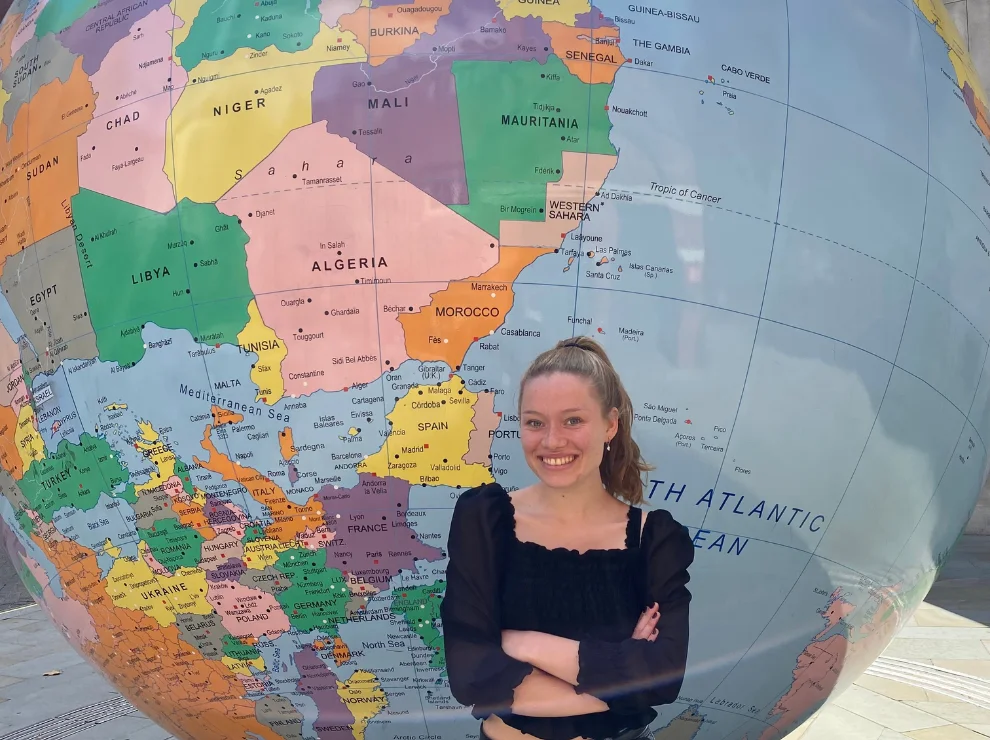 Eine junge Frau steht mit verschränkten Armen vor einer riesigen Skulptur einer Weltkugel und lächelt in die Kamera.