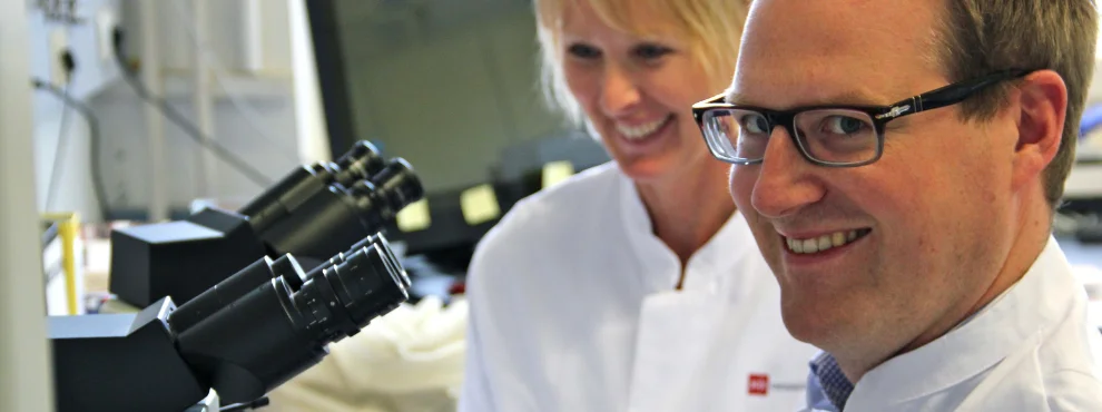 Ein Mann sitzt an einem Mikroskop und lächelt in die Kamera. Hinter ihm ist eine Frau zu sehen.
