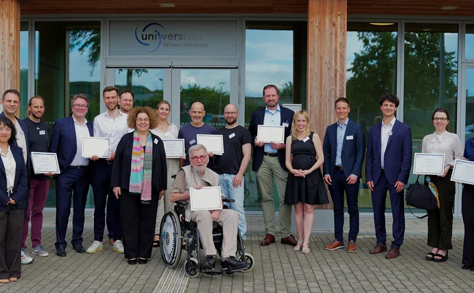 Bei einem Netzwerktreffen an der Universität Witten/Herdecke trafen sich Vertreter:innen von digitalen Vorreitern der Patient:innenversorgung in Nordrhein-Westfalen. (Foto: UW/H) Gruppenfoto vom ATLAS-Netzwerktreffen vor dem Eingang des Holzgebäudes der Uni Witten/Herdecke.