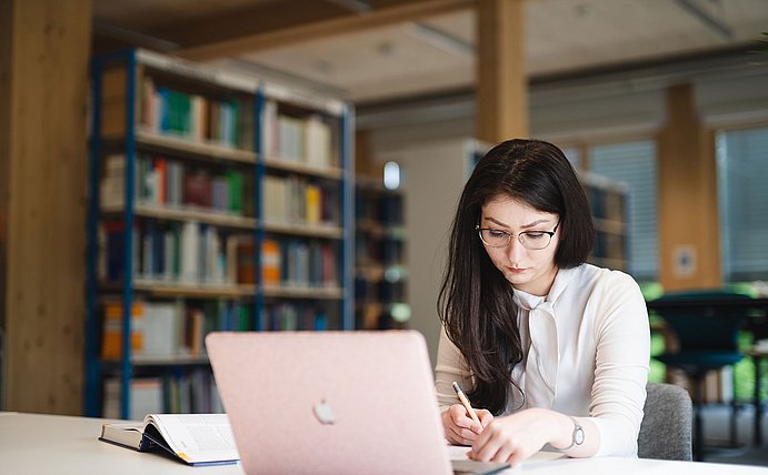 Eine Studentin sitzt an einem Schreibtisch und lernt. Vor ihr steht ein Laptop, daneben liegt ein aufgeschlagenes Buch. Im Hintergrund stehen Bücherregale.