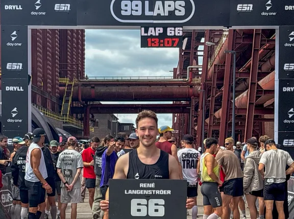 Beim Lauf-Event „99 Laps“ gehörte René zu 100 Teilnehmenden, die Runde für Runde gegeneinander antraten. René hielt 69 Runden durch und legte dabei 82,8 Kilometer zurück. (Foto: Privat) René Langenströer hält sein Laufergebnis auf einer Tafel fest und steht vor dem Zieleinlauf. Hinter ihm stehen weitere Läufer:innen.