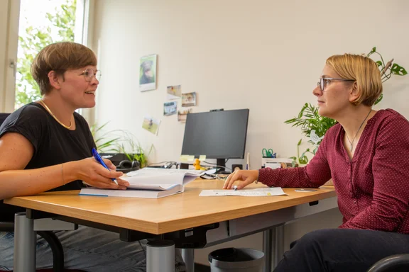 Dr. Sonja Knobbe und Prof. Dr. Magdalene Silberberger sitzen sich an einem Schreibtisch gegenüber.