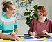 Zwei Frauen sitzen zusammen an einem Tisch und schauen sich an. Vor ihnen liegen verschiedene Poster.