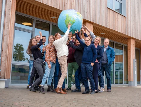 A group of people are standing in front of the entrance to the UW/H wooden building. With their arms outstretched, they are holding an inflatable globe aloft.