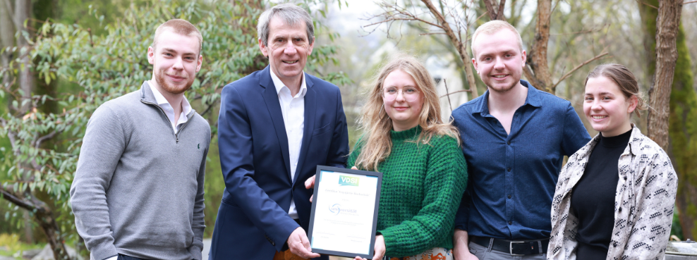 Fünf Personen stehen nebeneinander und lächeln in die Kamera. Zwei halten eine Urkunde hoch.