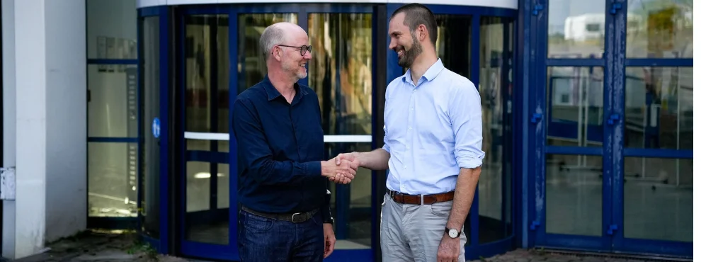 You can see the former dean, Prof. Dr. Dirk Sauerland, and the new dean, Prof. Dr. Nils-Christian Bormann, standing in front of the university's main building and shaking hands.