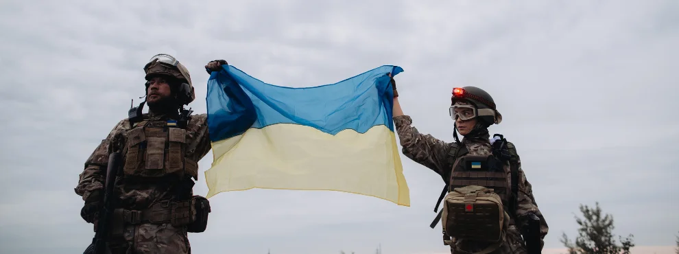 A male soldier on the left and a female soldier on the right are holding a Ukrainian flag in their hands.