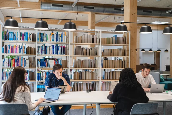 Co-Working-Space in der Bibliothek (Foto: UW/H | Johannes Buldmann) Vier Studierende sitzen an einem langen Tisch mit ihren Laptops und Büchern, um zu lernen.