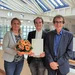 Drei Personen lächeln in die Kamera. Die Person in der Mitte hält eine Urkunde in den Händen. Die Person links überreicht der Person in der Mitte einen Blumenstrauß.