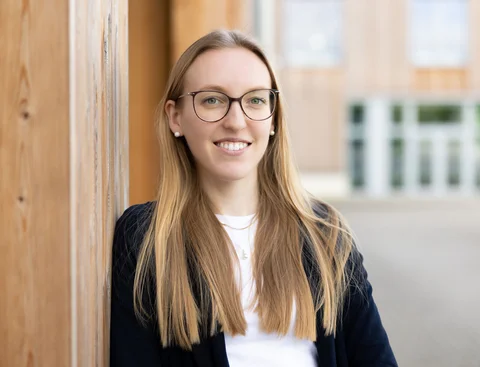 Bewirb dich für den Studiengang Psychologie (B. Sc.) an der UW/H. (Foto: UW/H | Michael Schwettmann) Eine Frau steht seitlich gelehnt an einer Gebäudewand aus Holz.
