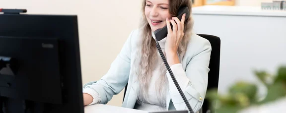 Junge Frau sitzt an einem Schreibtisch vor einem Computer und telefoniert.