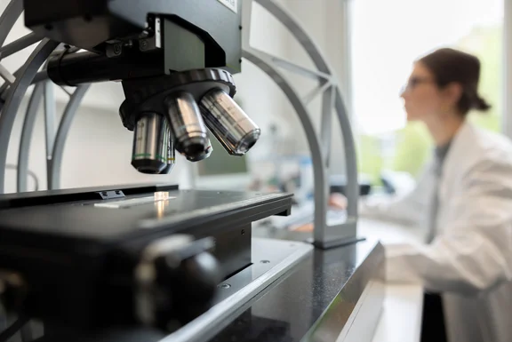 Close-up of a microscope in the foreground, a scientist at her desk in the background out of focus