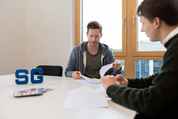 A student is sitting at a meeting table with an adviser. They are looking through contract documents together. On the table is a display stand from the StudierendenGesellschaft..