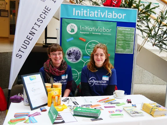 Emmi Mentzel und eine Kommilitonin sitzen an einem Infostand. Im Hintergrund sind eine Beachflag und eine Posterwand mit der Aufschrift "Initiativlabor".