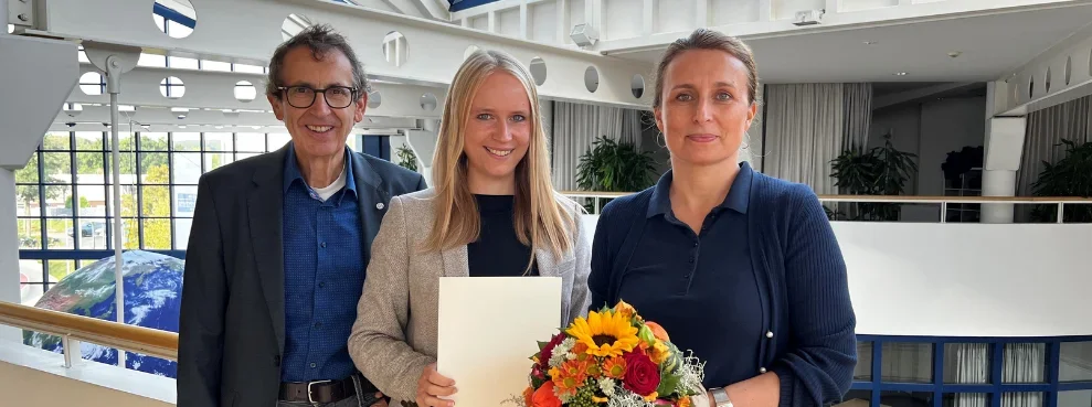 Drei Personen lächeln in die Kamera. Die Person in der Mitte hält eine Urkunde hoch.