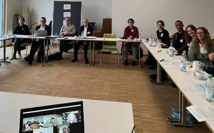 Eine Gruppe Menschen sitzt an U-förmig gestellten Tischen und schaut in die Kamera.