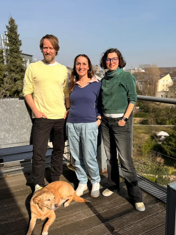 Zwei Frauen und ein Mann posieren nebeneinander für ein Bild. Zu ihren Füßen liegt ein Hund.