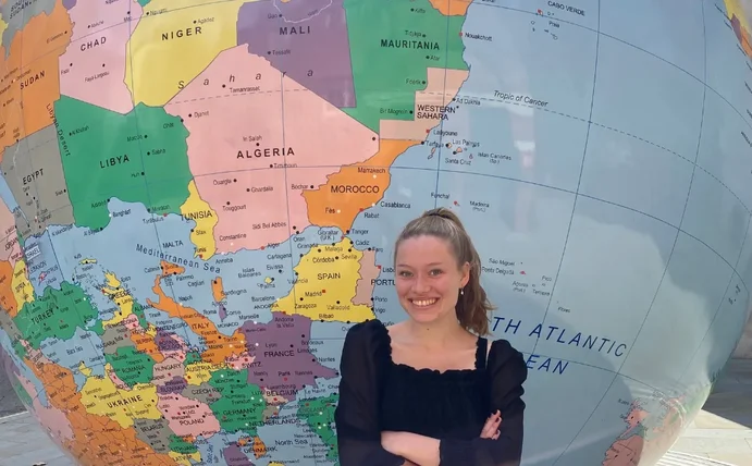 Eine junge Frau steht mit verschränkten Armen vor einer riesigen Skulptur einer Weltkugel und lächelt in die Kamera.