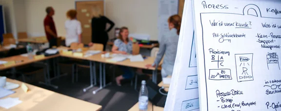 Die UW/H bietet zahlreiche Benefits, darunter ein umfassendes Weiterbildungsprogramm. (Foto: UW/H) Flipchart mit Grafiken im Vordergrund, im Hintergrund reden fünf Personen.