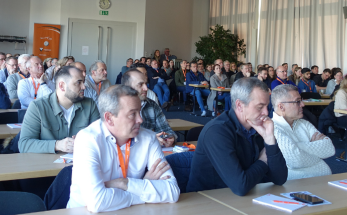 Many people sit at tables in a room, look ahead and listen.