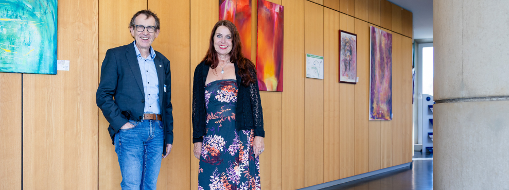 Prof. Dr. Stefan Zimmer begrüßt die Künstlerin Beate Braun mit ihren Werken in den Räumen der Zahnklinik der Universität Witten/Herdecke. (Foto: UW/H | Michael Schwettmann) EIn Mann und eine Frau stehen in einem Flur. An der Wand hinter ihnen hängen farbige Bilder.