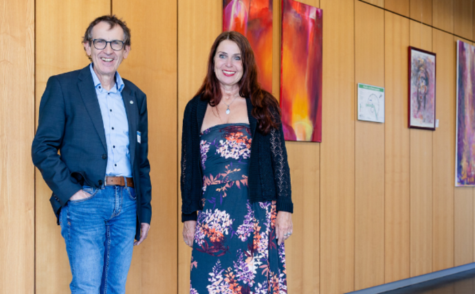 Prof. Dr. Stefan Zimmer begrüßt die Künstlerin Beate Braun mit ihren Werken in den Räumen der Zahnklinik der Universität Witten/Herdecke. (Foto: UW/H | Michael Schwettmann) EIn Mann und eine Frau stehen in einem Flur. An der Wand hinter ihnen hängen farbige Bilder.