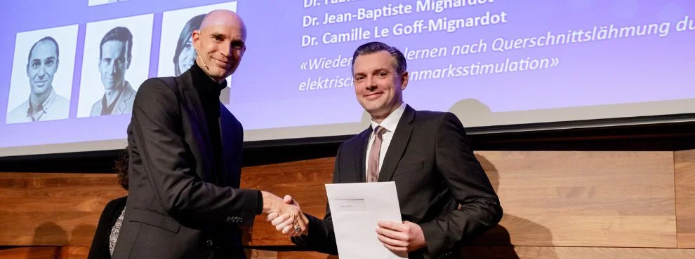 Zwei Männer schütteln sich auf einer Bühne die Hand und lächeln in die Kamera. Der rechte hält eine Urkunde in der Hand.