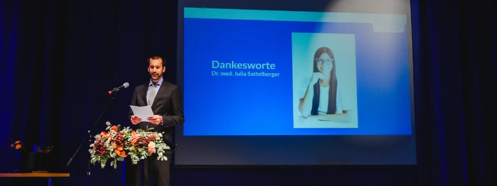 The laudator stands on the stage. In the background you can see the photo of the award winner on a screen.