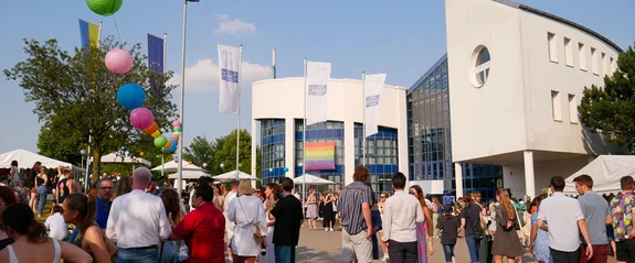 Eine Menschenmenge steht auf einem Campusvorplatz. Im Hintergrund ist das Campusgebäude. Entlang der Bäume ist eine bunte Lampion-Girlande gespannt.