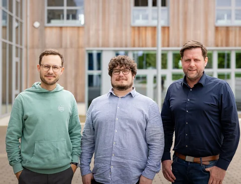 Das Team des Alumni-Management (Foto: UW/H | Michael Schwettmann) Ein Gruppenbild mit drei Männern vor dem Holzgebäude der Uni Witten/Herdecke.