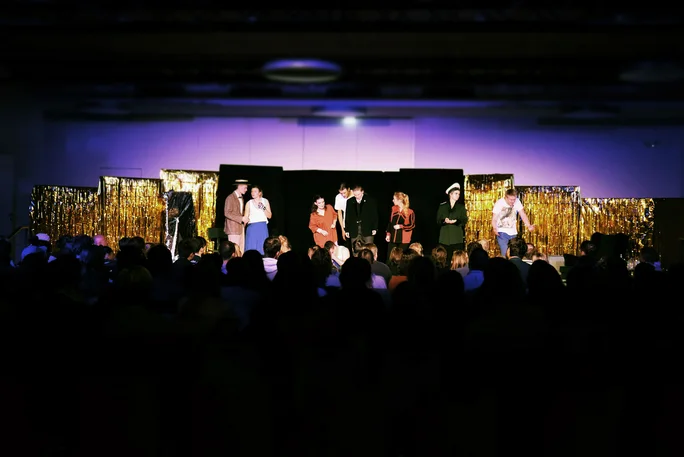 Im Theatersaal ist es sehr dunkel, nur die Bühne ist erleuchtet. Im Vordergrund sind die Silhouetten des Publikums zu erkennen, im Hintergrund stehen schauspielende Studierende in ihren Kostümen auf der Bühne.