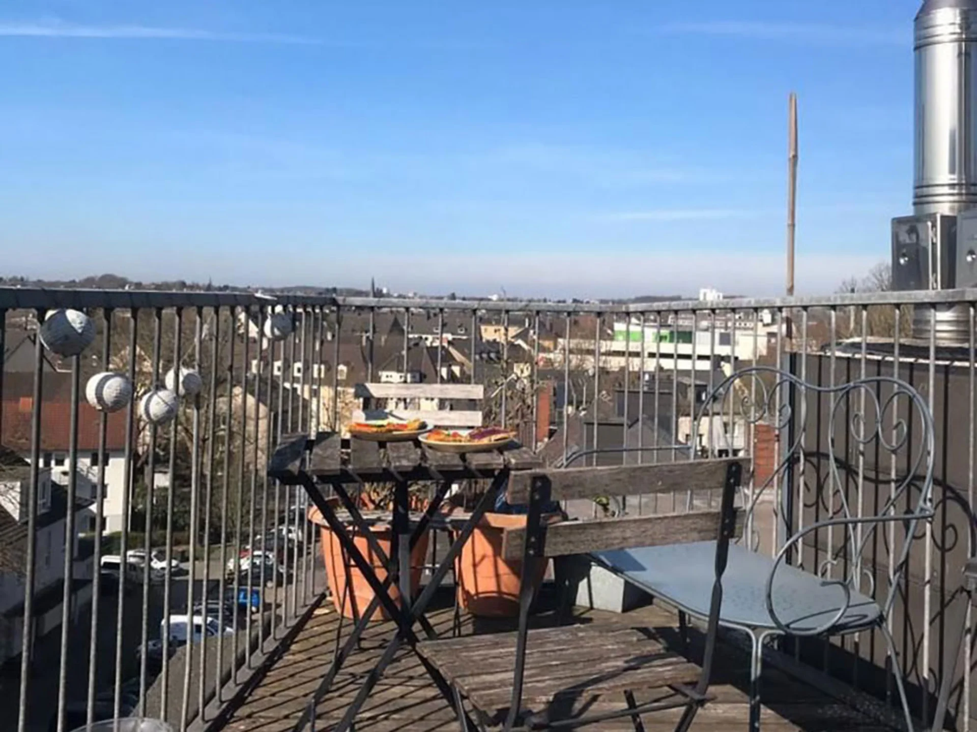 Roof terrace with a view over Witten (Photo: Private) On a small roof terrace there are two wooden folding chairs, a bench and a table. The terrace is surrounded by a metal railing. Half of the photograph shows a brilliant blue sky.