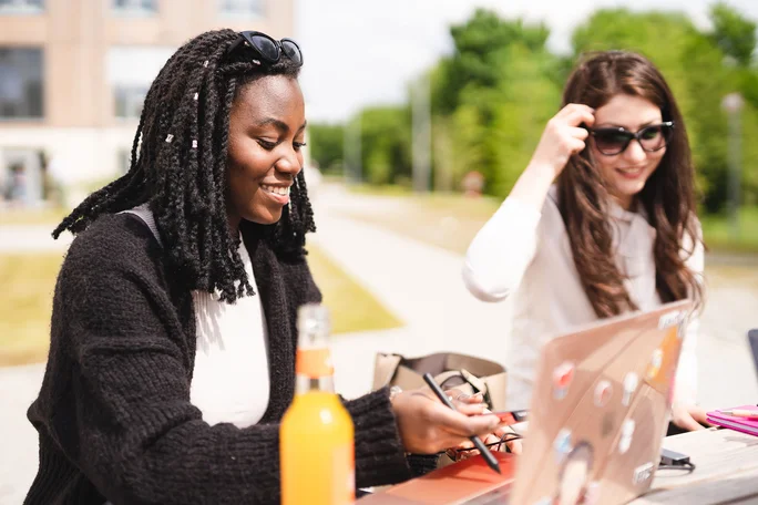 Lernen auf dem schönen Campus der Universität (Foto: UW/H | Johannes Buldmann) Zwei Studentinnen lernen mit Laptop auf dem Campus und bei Sonnenschein.
