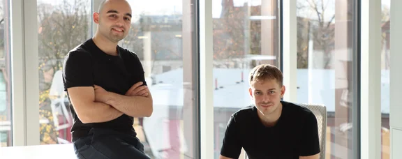Die beiden skulio-Gründer: Teoman Köse und Elias Perez (Foto: skulio) Foto von zwei Männern vor einer Fensterfront. Eine sitzt auf der Tischkante, der andere daneben auf einem Stuhl.