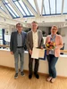 Drei Menschen stehen im Hauptgebäude der Uni W/H. In der Mitte steht Prof. Dr. Dr. Lars Bonitz, der mit einem Lächeln seine Berufungsurkunde in der Hand hält. 
