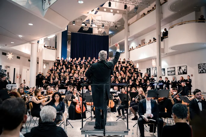 Christmas concert with the UW/H choir and orchestra. (Photo: UW/H | Daniela Arndt) In the foreground, the conductor looking towards the orchestra and choir. All the musicians are dressed in black. The main hall of the university is decorated with Christmas stars.