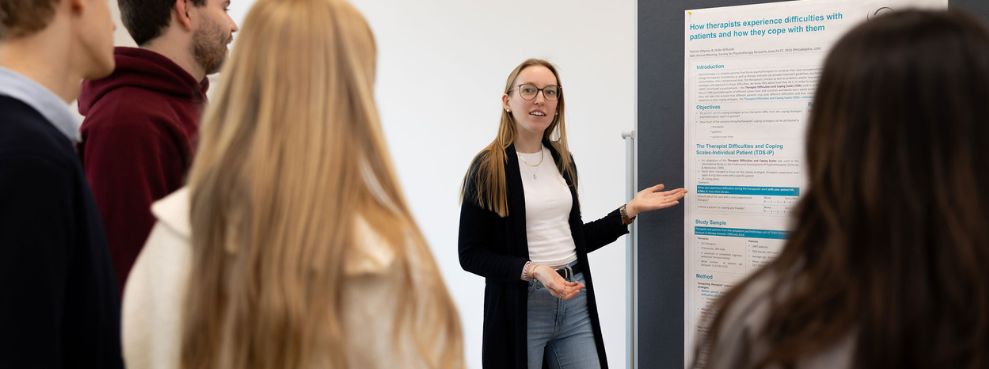A student points to a poster hanging on a metaplan wall and talks to other students.