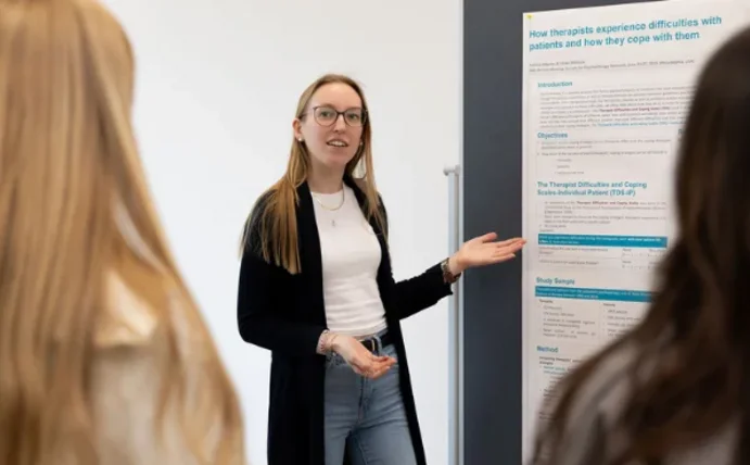 Eine Studentin zeigt auf ein Plakat, das an einer Metaplanwand hängt, und spricht dabei mit anderen Studierenden.