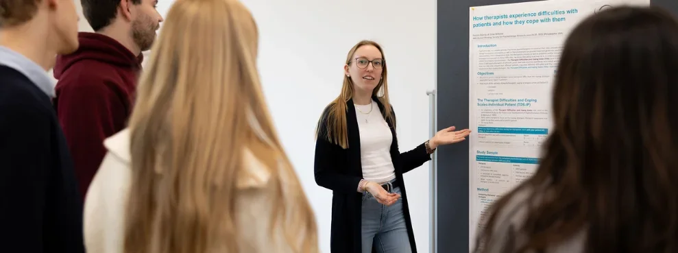 A student points to a poster hanging on a metaplan wall and talks to other students.