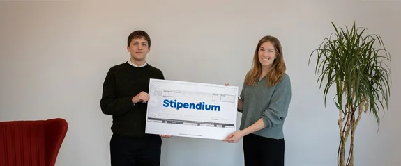 Zwei Studierende halten einen symbolischen, übergroßen Scheck mit der Aufschrift "Stipendium" in den Händen.