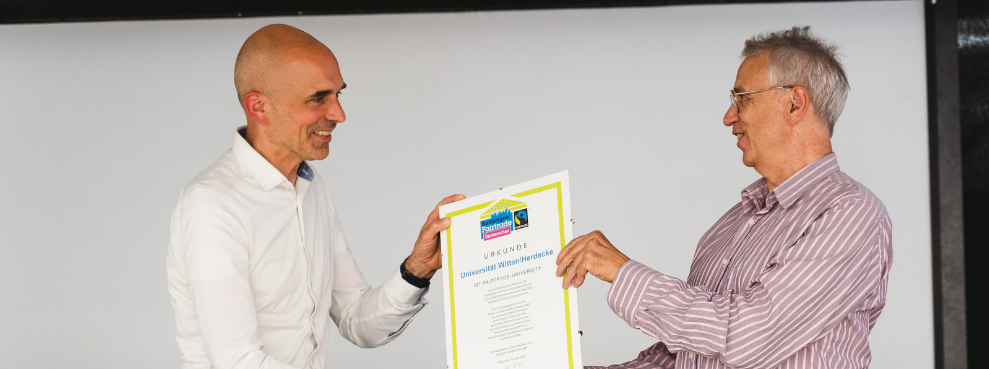 Dieter Overath, geschäftsführender Vorstand von Fairtrade Deutschland (rechts im Bild) übergibt UW/H-Vizepräsident Dr. Dirk Jakobs die Urkunde. (Foto: UW/H | Johannes Buldmann) Zwei Männer halten gemeinsam eine Urkunde fest.