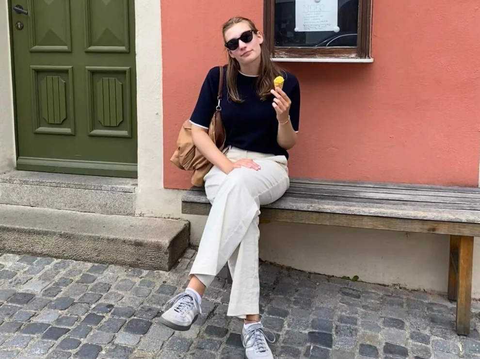 Pia Klee sitzt auf einer Holzbank vor einer roten Hauswand und isst ein Eis.