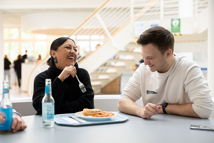 Zwei Studierende sitzen gemeinsam an einem Tisch in der Cafeteria und lachen. Vor ihnen steht ein Teller mit Essen sowie ein Getränk.